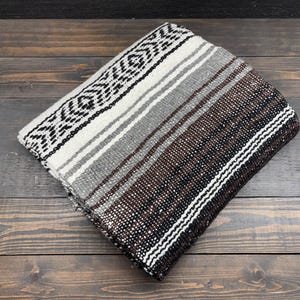 May include: A folded woven blanket featuring a repeating geometric pattern in black, white, gray, and dark brown. The textile displays a series of horizontal stripes and intricate diamond-like motifs across its surface, showcasing a textured weave.