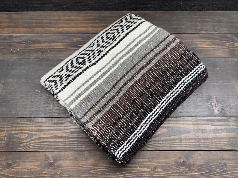May include: A folded woven blanket featuring a repeating geometric pattern in black, white, gray, and dark brown. The textile displays a series of horizontal stripes and intricate diamond-like motifs across its surface, showcasing a textured weave.