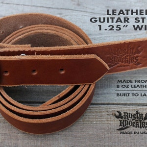 May include: Brown leather guitar strap with a silver buckle. The strap is 1.25 inches wide and is made from 8 ounce leather. The strap is labeled "Rusty Knuckles" and "Made in USA".