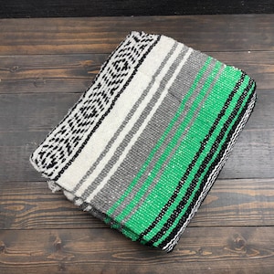 May include: A folded woven blanket featuring a vibrant green, white, gray, and black striped pattern. The textile displays a repeating geometric diamond design along one edge, transitioning into solid color bands of varying widths across the surface.