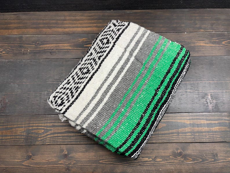 May include: A folded woven blanket featuring a vibrant green, white, gray, and black striped pattern. The textile displays a repeating geometric diamond design along one edge, transitioning into solid color bands of varying widths across the surface.