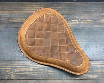 May include: Brown leather motorcycle seat with a diamond-stitched pattern. The seat has a rounded shape and a distressed finish, suggesting a vintage or custom design. The stitching is a lighter color, providing contrast.