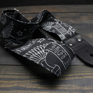 May include: Black guitar strap with white tattoo-style designs, including a banner that reads "LOVE", stars, and a skull. The strap has black leather ends and metal rivets. The strap is laying on a dark wooden surface.