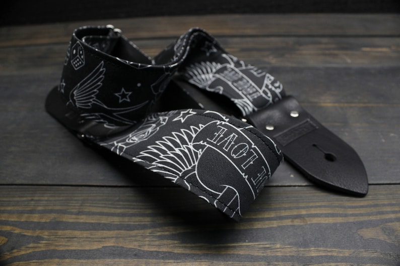 May include: Black guitar strap with white tattoo-style designs, including a banner that reads "LOVE", stars, and a skull. The strap has black leather ends and metal rivets. The strap is laying on a dark wooden surface.
