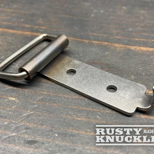 May include: A metal belt buckle with a rectangular frame and a hook. The buckle is made of a brushed metal material and has two holes on the plate. The text "RUSTY KNUCKLES MADE IN USA" is printed on the bottom.