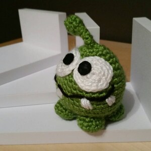 May include: Green crocheted plush toy with large white eyes and a toothy grin. The toy is sitting on a white shelf.