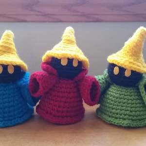 May include: Three crocheted plushies in the shape of a black mage from the Final Fantasy video game series. The plushies are wearing yellow pointed hats and have black eyes. The plushies are wearing blue, red, and green robes.