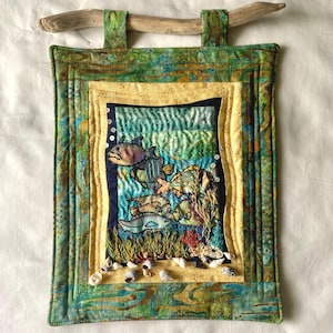 May include: A rectangular fabric wall hanging featuring a quilted underwater scene with embroidered fish and seaweed. The piece is framed by green and yellow patterned fabric borders and is suspended from a piece of driftwood with small seashells.