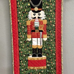 May include: A red, white, and black nutcracker doll stands on a red and gold drum. The doll is holding a silver sword and has a white beard and mustache. The background is a green fabric with a Christmas tree pattern.