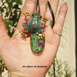 Puede incluir: Un collar con colgante de rana. El colgante presenta una rana enjoyada verde y rosa sobre una piedra verde y roja. El colgante cuelga de un cordón negro. El texto "les bijoux de Guylaine" está en la parte inferior.