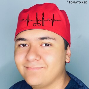 Può includere: Un berretto rosso da scrub con un grafico di battito cardiaco nero e strumenti chirurgici. Il testo "*TOMATO RED" è visibile nell'angolo in alto a destra dell'immagine.