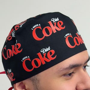 May include: Black surgical cap with a repeating red and white "Diet Coke" logo pattern. The cap has a red tie in the back. The text "I need a Diet Coke" is also printed on the cap.
