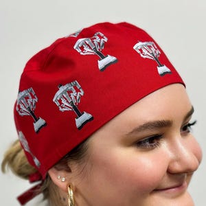 Könnte beinhalten: Eine rote OP-Haube mit einem sich wiederholenden Muster aus silbernen und schwarzen Trophäen-Designs. Die Haube ist hinten gebunden. Die Haube besteht aus einem weichen, atmungsaktiven Stoff und wird von einer Person getragen.