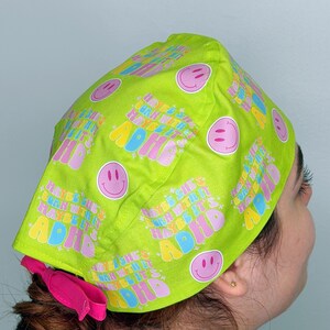 May include: A bright green scrub cap featuring a repeating pattern of smiley faces and the text "Maybe she's born with it, maybe it's ADHD" in a colourful, retro font. A pink scrunchie is visible at the back.