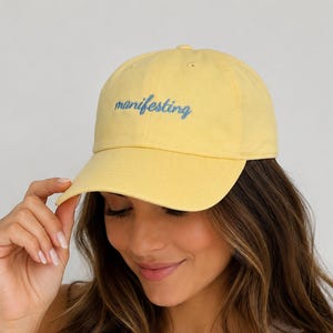 May include: A pale yellow baseball cap featuring the word manifesting embroidered in a cursive blue script font across the front panel. The cap has a structured crown, a curved brim, and eyelets for ventilation on the top panels of the hat.