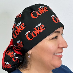 May include: Black scrub cap with a repeating pattern of the words "Diet Coke" and "I Need a Coke" in red and white. The cap has a red ribbon detail. The cap is worn by a person.