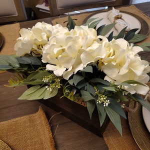 May include: A wooden box filled with white hydrangeas and green eucalyptus leaves, arranged as a centerpiece on a table with burlap placemats.