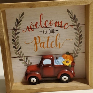 May include: A wooden framed sign with a red truck carrying pumpkins and a sunflower. The sign says "Welcome to our Patch".