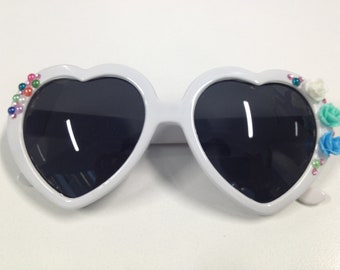 May include: White heart-shaped sunglasses with black lenses and colorful embellishments, including beads and flowers.