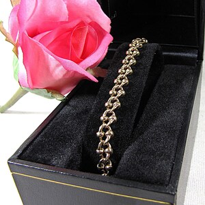 May include: A gold-toned bracelet with a unique linked design rests in a black jewelry box. A pink rose is positioned next to the box. The bracelet is displayed on a black velvet insert.