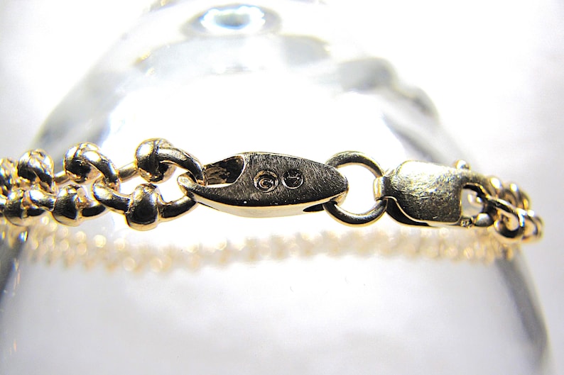 May include: Close-up of a gold-colored bracelet with a decorative chain design. The bracelet features a secure clasp and is displayed against a blurred background. The jewelry piece is likely made of gold.