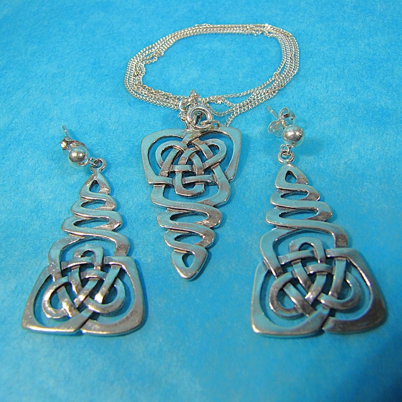 May include: A silver-coloured jewellery set with a Celtic knot design. The set includes a pendant necklace and matching earrings. The pendant and earrings have an openwork design with intricate knot patterns. The jewellery is displayed on a blue background.