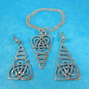 May include: A silver Celtic knot jewellery set comprising a pendant necklace and matching earrings. The pendant and earrings feature an open, flowing design with intricate knotwork. The jewellery is displayed against a solid blue background.