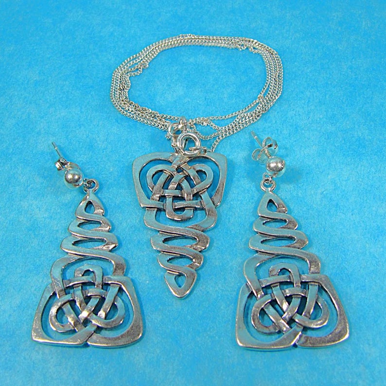 May include: A silver Celtic knot jewellery set comprising a pendant necklace and matching earrings. The pendant and earrings feature an open, flowing design with intricate knotwork. The jewellery is displayed against a solid blue background.