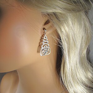 May include: Silver Celtic knot earrings. The earrings have a post-back fastening and a dangling design with intricate knotwork. The earrings are displayed on a mannequin, highlighting their design and style. The earrings are a classic accessory.