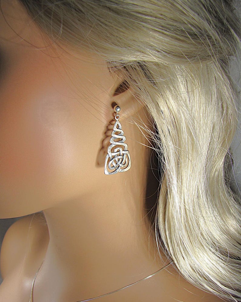 May include: Silver Celtic knot earrings. The earrings have a post-back fastening and a dangling design with intricate knotwork. The earrings are displayed on a mannequin, highlighting their design and style. The earrings are a classic accessory.