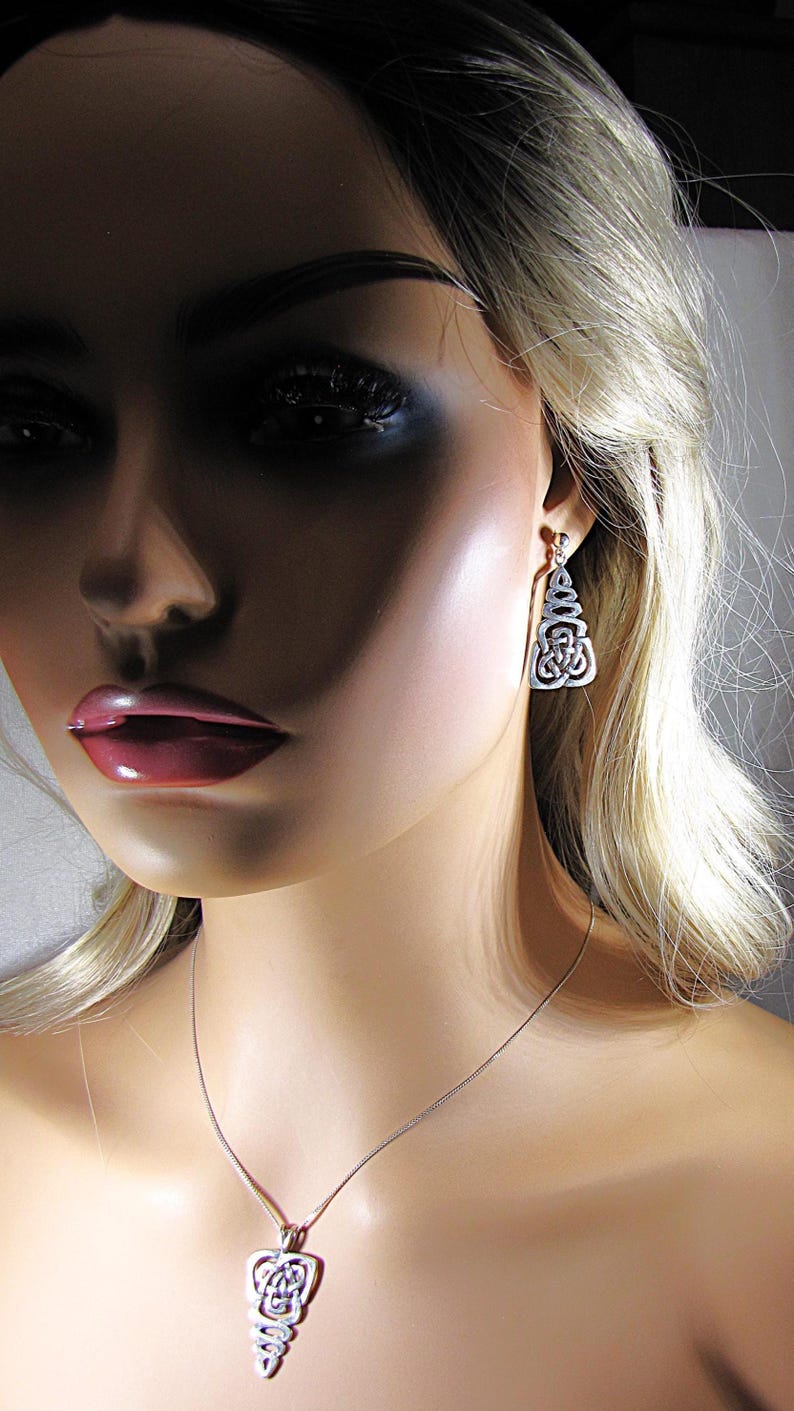 May include: Silver-toned pendant necklace and matching earrings. The pendant and earrings feature an openwork design with a Celtic knot motif. The jewelry is displayed on a mannequin with blonde hair and a fair complexion.