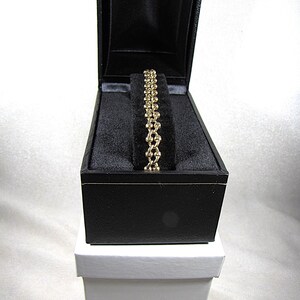 May include: A gold-colored bracelet with a chain-link design rests inside a black jewelry box. The box is open, revealing the bracelet against a black velvet interior. The box sits atop a white box.
