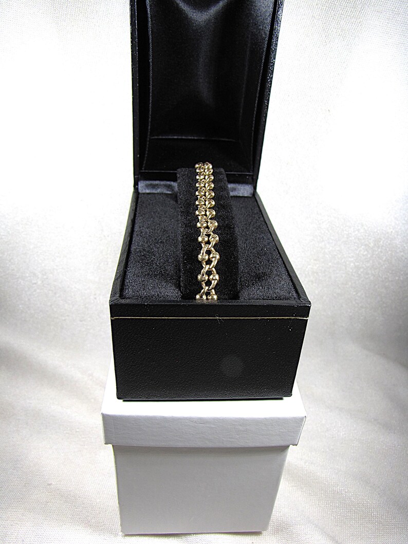 May include: A gold-colored bracelet with a chain-link design rests inside a black jewelry box. The box is open, revealing the bracelet against a black velvet interior. The box sits atop a white box.