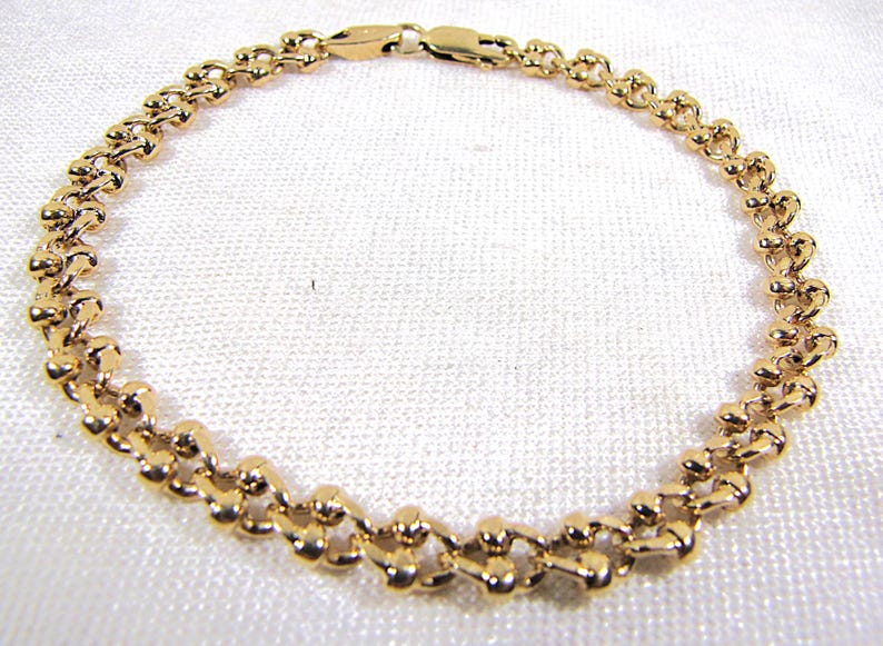 May include: A gold-colored bracelet with a delicate, linked design. The bracelet features a series of small, rounded links connected to form a flexible chain. The clasp is also gold-toned. The bracelet is displayed on a white surface.
