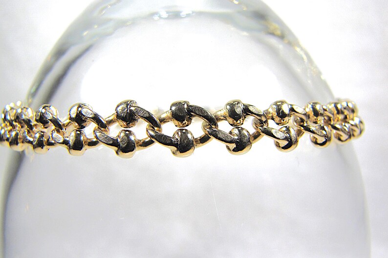 May include: A gold-colored bracelet with a chain-link design. The links are oval-shaped and connected in a repeating pattern. The bracelet is displayed against a blurred, light background.