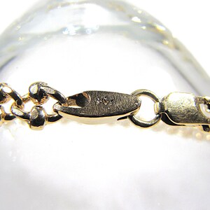 May include: Close-up of a gold-colored bracelet clasp and chain. The chain links are small and rounded, with a textured surface. The clasp is a lobster claw style, with a small ring for attachment.