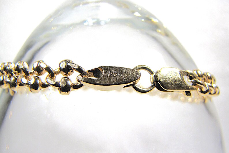 May include: Close-up of a gold-colored bracelet clasp and chain. The chain links are small and rounded, with a textured surface. The clasp is a lobster claw style, with a small ring for attachment.