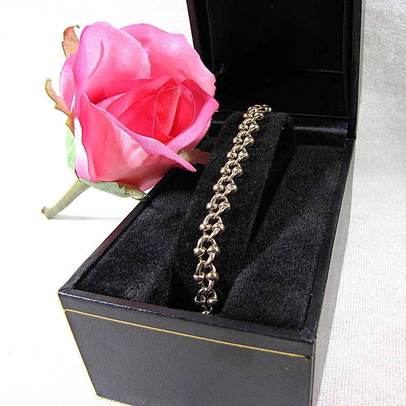 May include: A gold-toned bracelet with a unique linked design rests in a black jewelry box. A pink rose is positioned next to the box. The bracelet is displayed on a black velvet insert.
