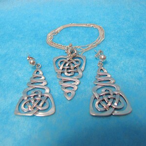 May include: A silver pendant necklace and matching earrings set with a Celtic knot design. The jewellery is displayed on a blue background. The necklace has a delicate chain. The earrings are drop style.