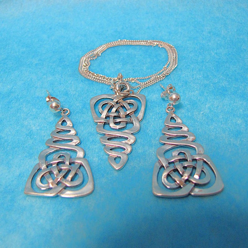 May include: A silver pendant necklace and matching earrings set with a Celtic knot design. The jewellery is displayed on a blue background. The necklace has a delicate chain. The earrings are drop style.