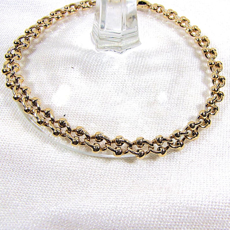 May include: A gold-colored bracelet with a double-strand chain design. The bracelet features small, round beads connected by delicate links, creating a textured and elegant appearance. The jewelry is displayed on a clear glass surface.