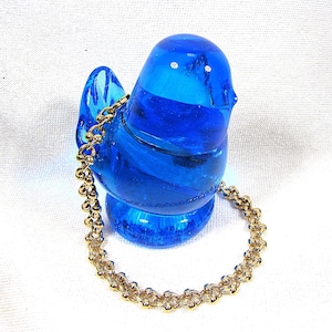 May include: A vibrant blue glass bird figurine with a gold-colored chain. The bird has a rounded body, a small tail, and a detailed head. The chain is draped around the bird, adding a decorative element. The figurine is set against a white background.