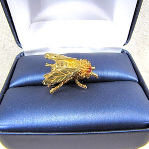 May include: A gold-colored bee brooch with intricate detailing, including textured wings and a red gemstone eye, rests on a blue satin cushion inside a dark blue jewelry box. The brooch is a decorative accessory.