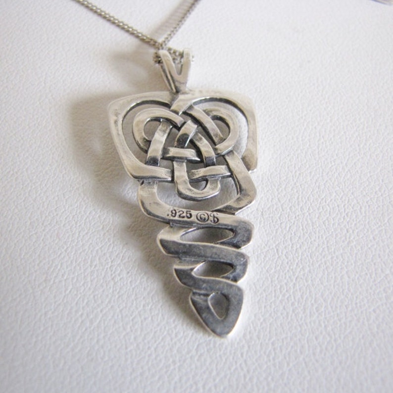 May include: A silver pendant necklace with a Celtic knot design. The pendant has a rectangular top with interwoven lines and a spiraling bottom. The necklace is on a silver chain and is marked with the number .925.