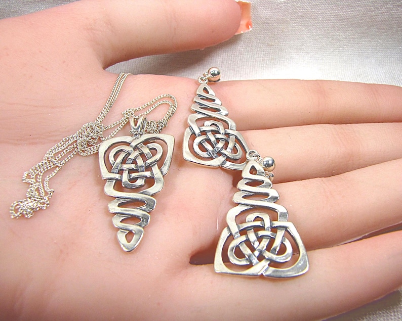 May include: A silver pendant necklace and matching earrings set. The pendant and earrings feature a Celtic knot design with a spiral base. The jewellery is displayed on a light background.