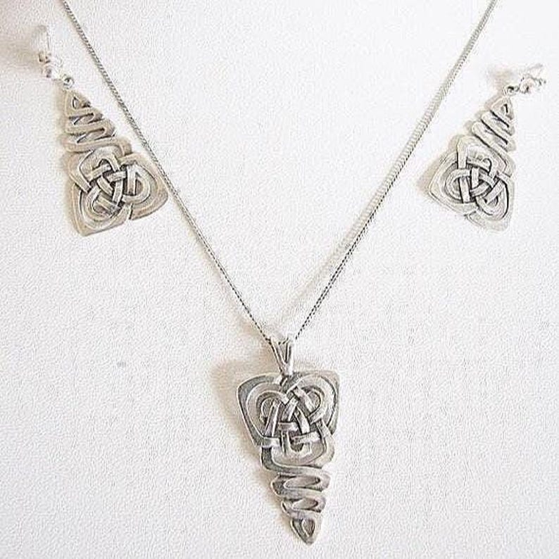May include: A silver-coloured jewellery set with a Celtic knot design. The set includes a pendant necklace and matching earrings. The pendant and earrings have a detailed, openwork design, and the necklace has a delicate chain.