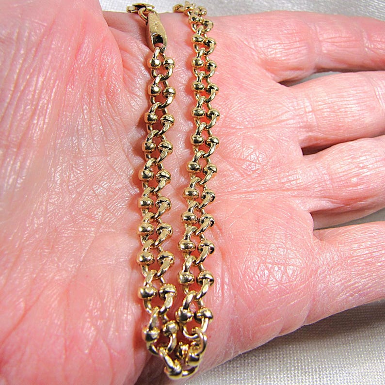 May include: A gold-colored chain bracelet with a unique beaded design. The bracelet is composed of small, connected beads, creating a textured and eye-catching accessory. The clasp is visible at the top. The bracelet is shown on a person's hand.