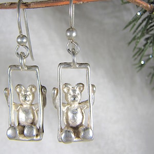 May include: A pair of silver-toned drop earrings featuring a three-dimensional teddy bear figure seated within a rectangular frame. Each bear has raised arms and is suspended from a simple silver-toned hook wire with a small spherical bead accent.