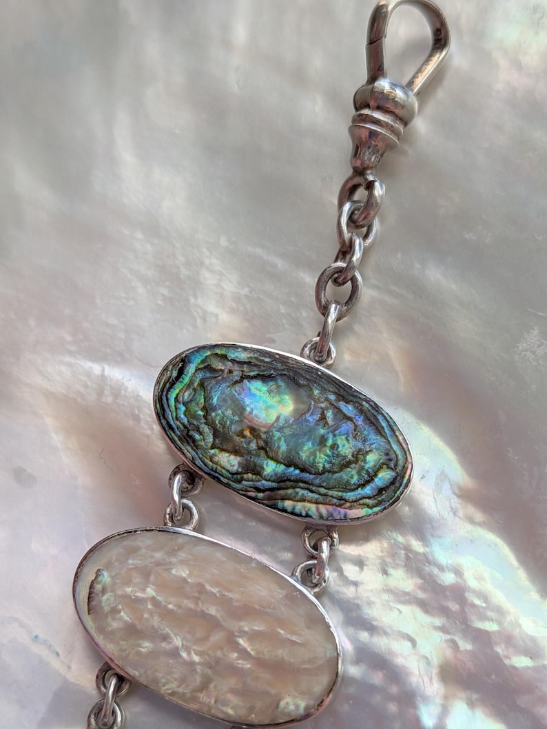 May include: A silver chain with a clasp and two oval-shaped abalone shell charms. The charms are iridescent with shades of green, blue, and purple.