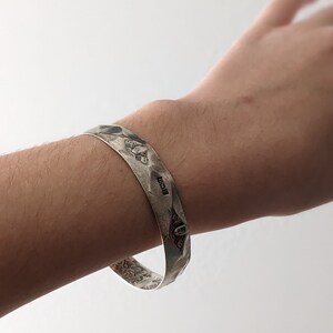 May include: A silver bracelet with a hammered finish and intricate engraved designs. The bracelet has a hallmark that reads "925".